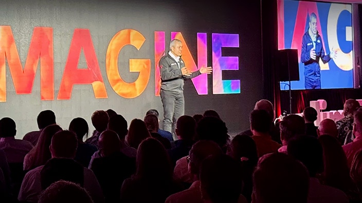 Nitin Tandon presents on stage in front of a colorful “IMAGINE” backdrop, with a live video feed displayed on a large screen and attendees seated in the audience.