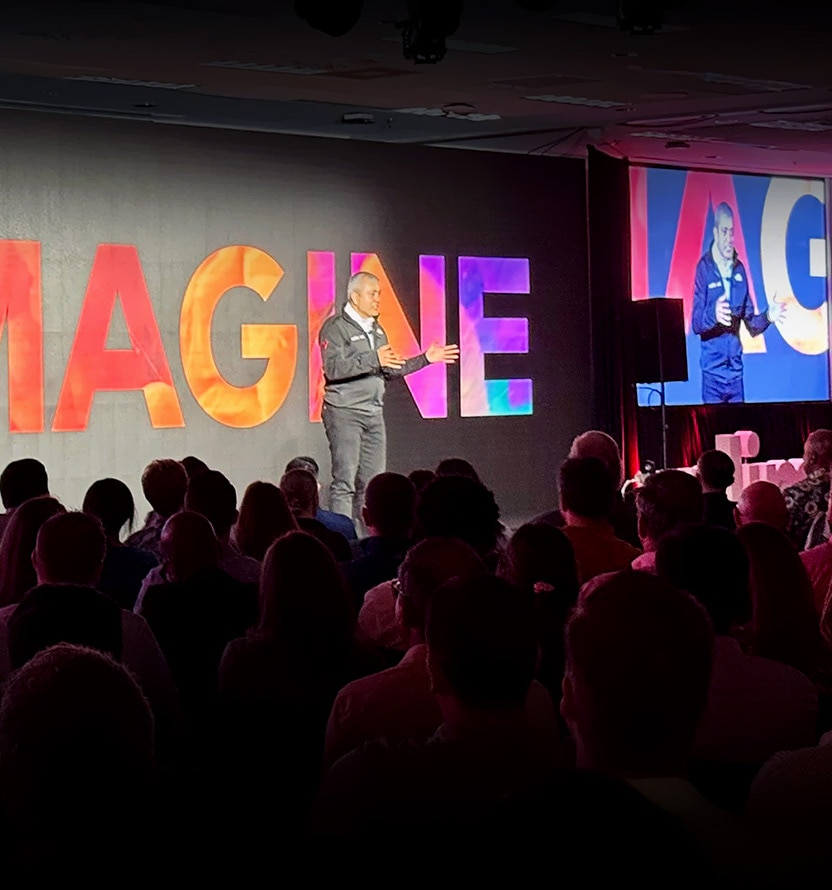 Nitin Tandon presents on stage in front of a colorful “IMAGINE” backdrop, with a live video feed displayed on a large screen and attendees seated in the audience.