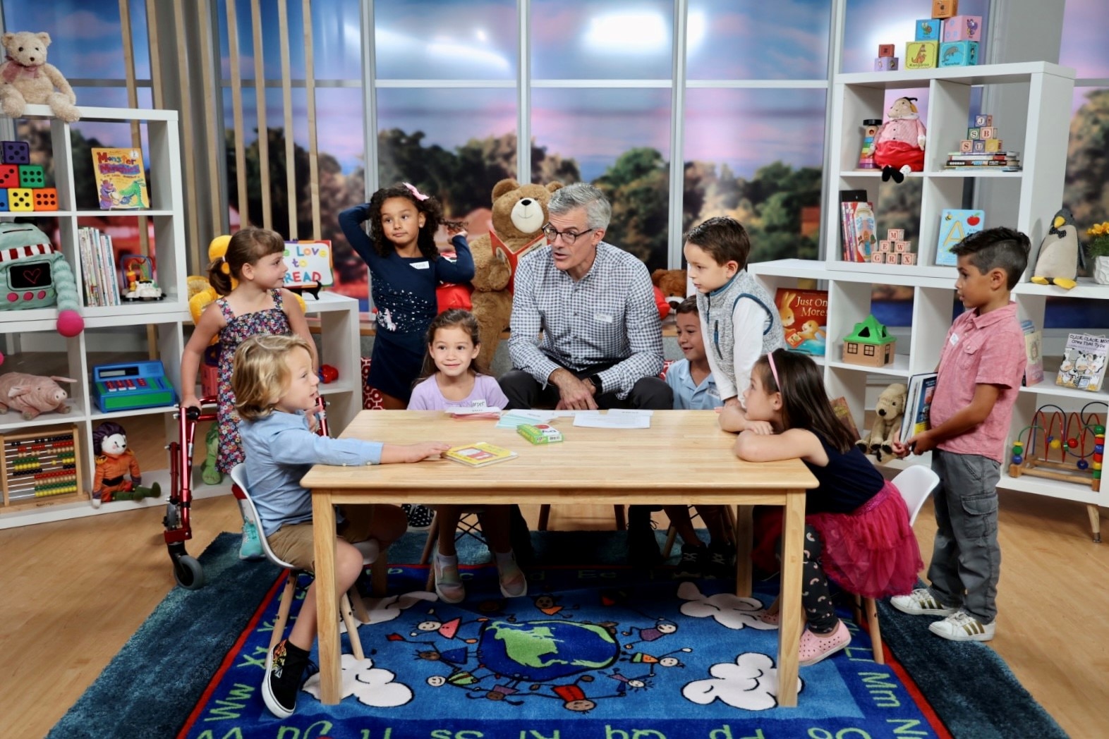 Vanguard CEO Tim Buckley sits on a chair behind a child-size wooden table in a brightly decorated room filled with books and children’s toys. Four children, 4 to 6 years old, are seated in small chairs at the table and four more children are standing. The children listen as Tim speaks with a boy seated at far left. 