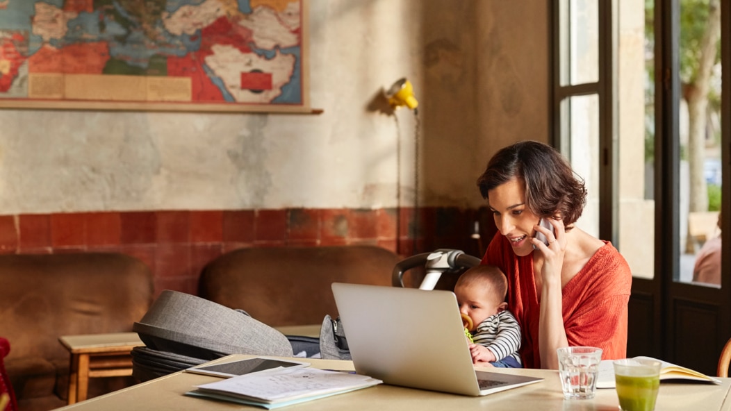 A person on the phone holding a young child and looking at a laptop computer. 