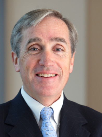 John J. Brennan is shown smiling. He has gray hair and is wearing a suit, dress shirt, and tie.