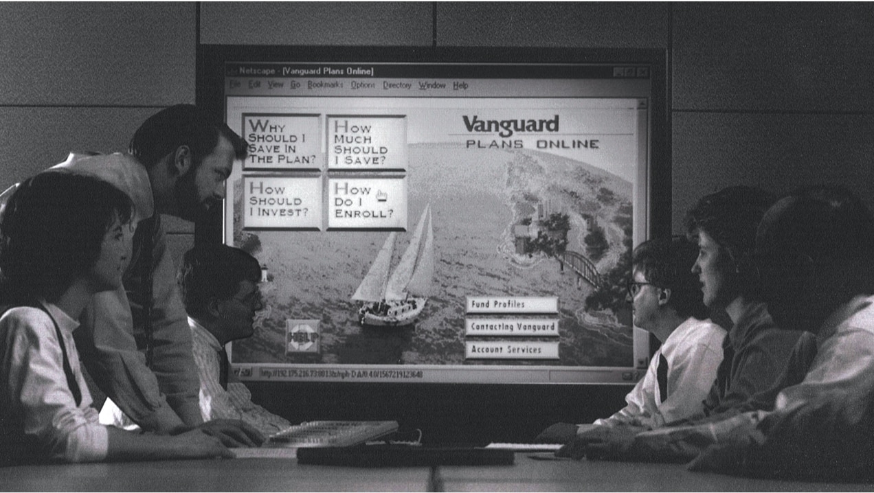 Four men and two women meet around a conference table. They are looking at a large screen showing a version of vanguard.com for investors in employer plans. The page features an image of a ship sailing toward a harbor and questions about why one should save, how much to save, how to invest, and how to enroll that link to answers and more information.