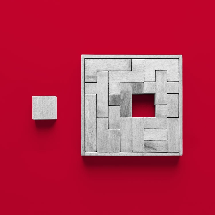 Square wooden puzzle with interlocking rectangular pieces forming a frame around a central square hole, placed on a bright red background. A single wooden block lies to the left of the puzzle.