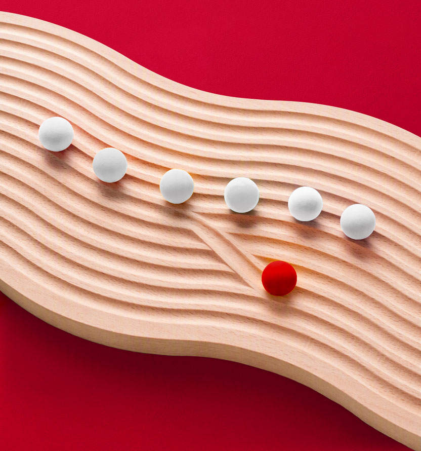 White spheres follow a winding path across a textured wooden surface, while a single red sphere diverges onto a separate route against a red background.