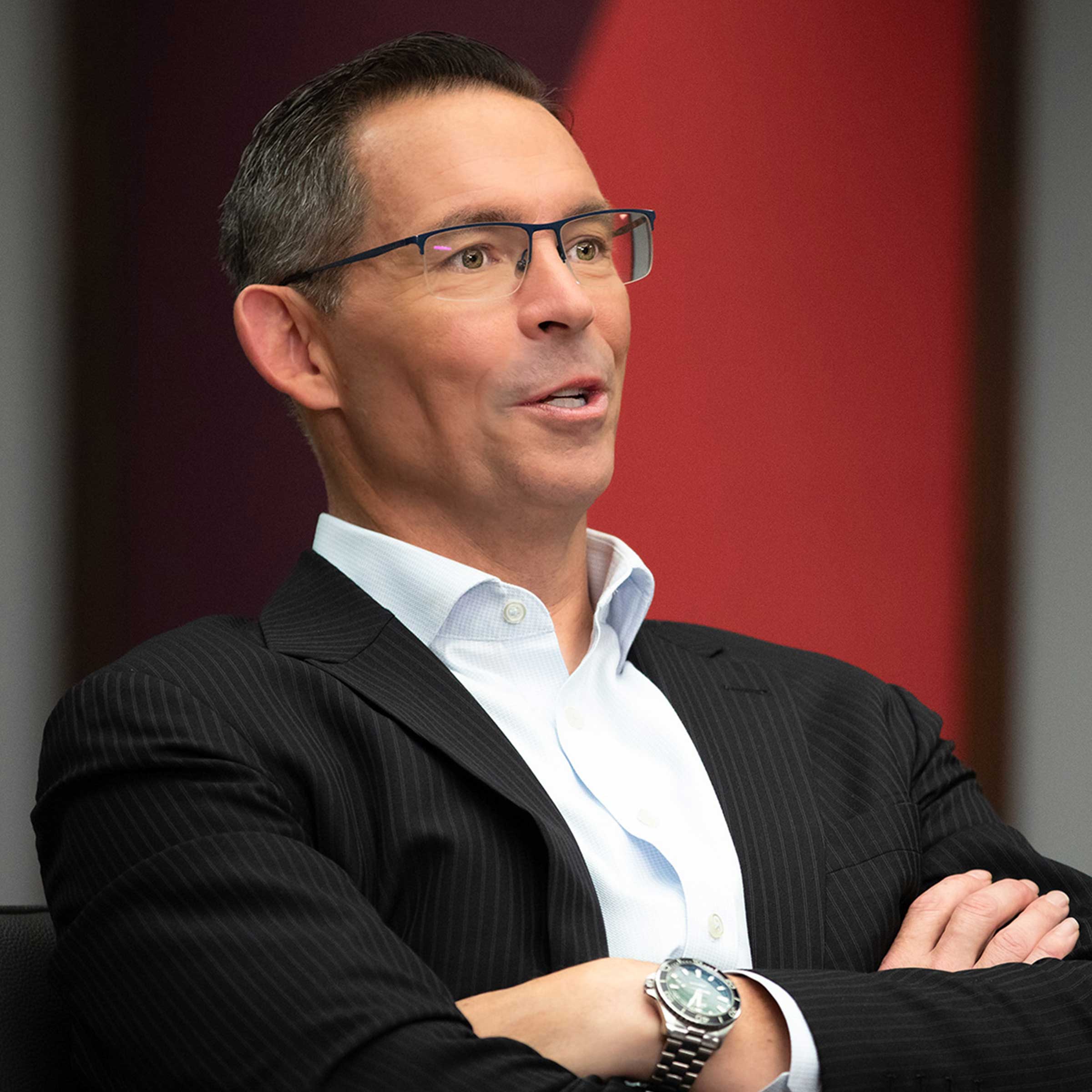 Joe Davis speaking into a microphone while seated with arms crossed, wearing a dark suit jacket and glasses.