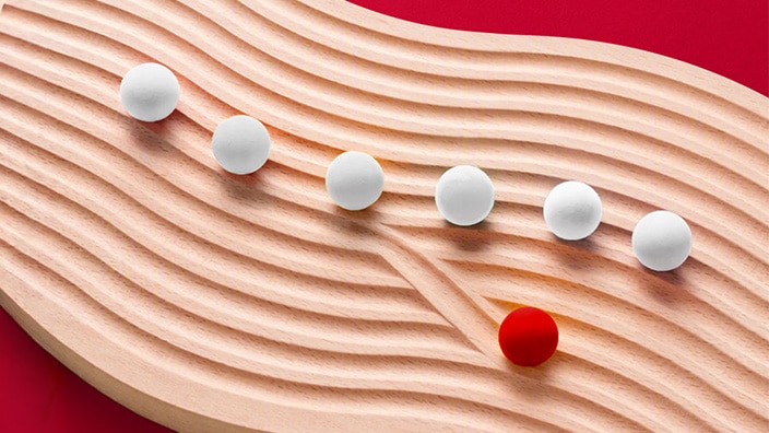 White spheres follow a winding path across a textured wooden surface, while a single red sphere diverges onto a separate route against a red background.