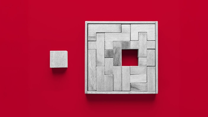 A minimalist image showing a small gray cube placed to the left of a larger square frame filled with interlocking gray rectangular blocks. The larger frame has a square hole in its center, and both objects sit on a bright red background, creating strong contrast.