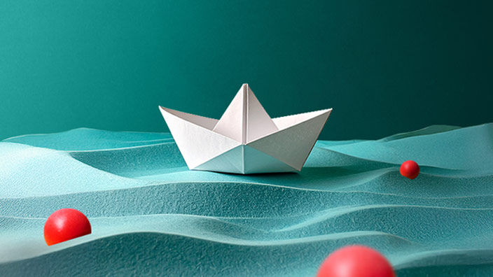 Paper sail boat floating on water made of blue sand with colored red balls floating around it