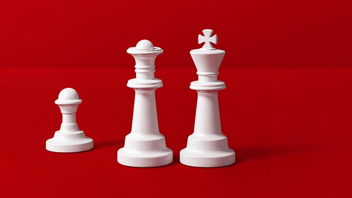 Three white chess pieces stand against a bright red background: a pawn on the left, a queen in the center, and a king on the right. The pieces are arranged in ascending height, with the king as the tallest piece, creating a simple, high-contrast composition.